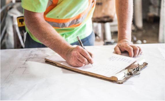 A man in a safety vest writing on a clipboard at a construction site.